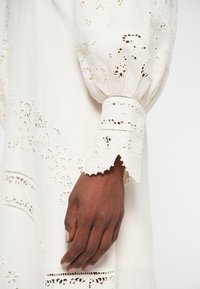 Robe blanche avec des accents en dentelle, dotée de manches bouffantes et d'une broderie finement détaillée, tissu texturé avec des motifs découpés.