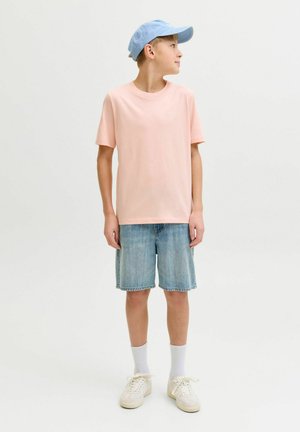 Dreng iført en lyseblå kasket, lyserød T-shirt, cowboyshorts, hvide sokker og sneakers, der står og ser til siden på en ensfarvet baggrund.