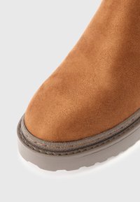 Τα λευκά σουέτ μποτάκια μέχρι τον αστράγαλο με στρογγυλή μύτη, αντίθετη γκρίζα καουτσούκ σόλα και ορατό ράψιμο κατά μήκος της άκρης. Μαλακή υφή και ανθεκτική σχεδίαση.