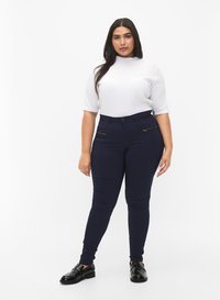 Frau steht mit einem weißen kurzärmeligen Oberteil, dunkelblauen Slim-Fit-Hosen mit Reißverschlusstaschen und schwarzen Slippers vor einem weißen Hintergrund.