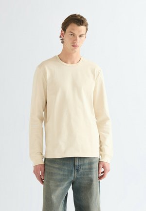 Giovane uomo in piedi, indossa una camicia beige a maniche lunghe e jeans blu scoloriti contro uno sfondo chiaro e uniforme.