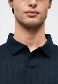 Polo bleu marine à texture côtelée, avec un col plié et une petite patte de boutonnage. Vue en gros plan du décolleté et de la partie supérieure de la poitrine.