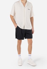 Man die een wit kort mouw overhemd met knopen draagt, zwarte korte broek, witte sokken en witte sneakers, staand met handen in de zakken.