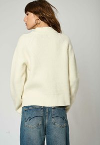 Pull crème texturé avec un col montant, des manches surdimensionnées et un ourlet côtelé, associé à un jean en denim bleu avec des poches arrière.
