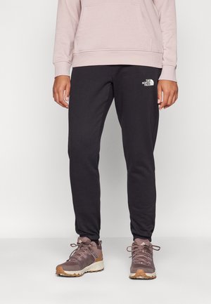 Osoba ubrana w czarne spodnie dresowe North Face i liliowe buty trekkingowe stoi na białym tle.