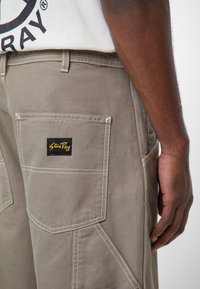 Beige cargo kalhoty s bílým šitím a bočními kapsami. Obsahují černou a žlutou nášivku. Vyrobeny z odolné látky, která zdůrazňuje strukturovaný design.