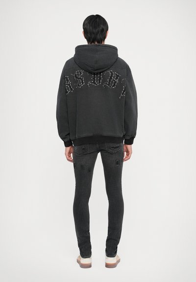 Černá oversized mikina s stylizovaným písmem na zádech, kombinovaná s černými slim-fit džíny s distressed detaily a manžetou na spodním okraji.