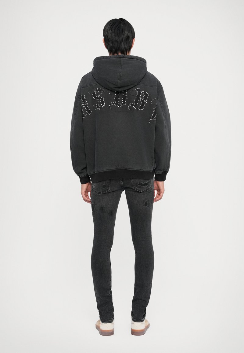 Čierna oversized mikina s kovovými nápismi na chrbte, v kombinácii s čiernymi slim-fit džínsami so zničenými detailmi a manžetou.