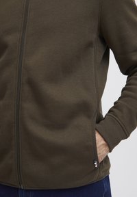 Temno zelen zip-up kapucar z gladko teksturo, stranskima žepoma in rebrastimi manšetami. Ima logotipne akcentne barve rdeče, bele in modre na robu.