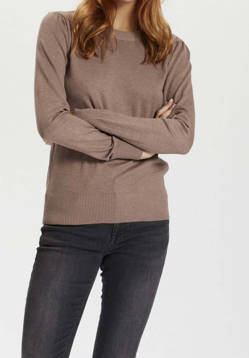 Ljusbrun stickad tröja med långa ärmar, ribbade manschetter och nederkant, med en mjuk textur och en klassisk rund halsringning. Bärs med svarta jeans.