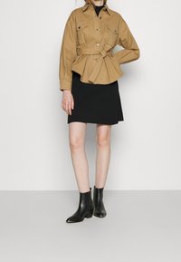 Veste beige à ceinture avec fermeture par boutons, deux poches poitrine, associée à une mini-jupe noire à côtes et des bottines noires.