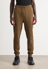 Man wearing brown jogger pants with drawstring waist and red patch logo on thigh, paired with black sneakers and black top.