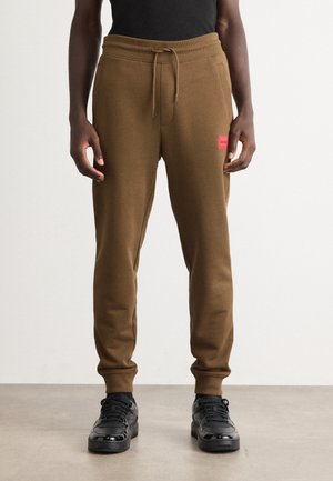 Mann med brune joggebukser med snor i livet og rødt logo på låret, sammen med svarte joggesko og svart topp.