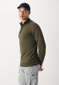 Pull à col zippé olive avec manches longues, ourlet côtelé et coutures aux épaules. Pantalons cargo gris avec poches latérales. Casquette noire portée.