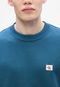 Felpa in cotone blu con colletto rotondo a coste. Presenta una piccola etichetta con logo bianco e scritta rossa nella parte sinistra del petto.