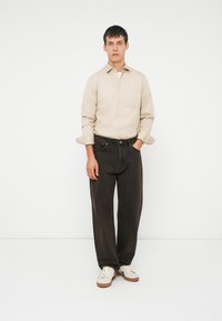 Μπεζ πουκάμισο με κουμπιά, σκούρα τζιν παντελόνια και λευκά αθλητικά παπούτσια. Το πουκάμισο έχει γυρισμένα μανίκια; το παντελόνι είναι σε άνετη γραμμή με ελαφριά υφή.