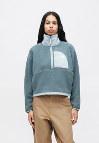 Άτομο με μακριά σκούρα μαλλιά, ντυμένο με ανοιχτό μπλε φλις πουλόβερ με φερμουάρ και μπεζ παντελόνι, που στέκεται μπροστά σε λευκό απλό φόντο.