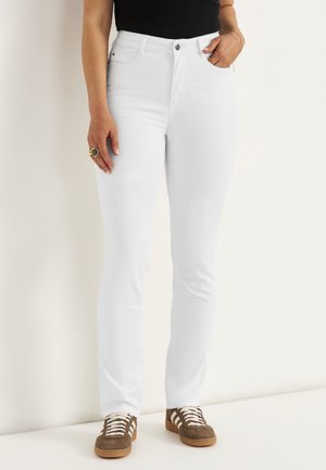 Femme portant un jean blanc ajusté, un haut noir, des baskets marron avec des lacets blancs, debout contre un mur blanc, les mains le long du corps.