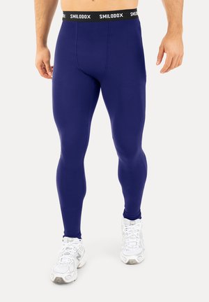 Mand iført marineblå leggings i fuld længde med en sort linning mærket "SMILODOX" og hvide sportssko.