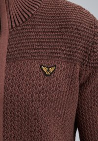 Sweter dziany w ciemnym brązie z teksturowanym wzorem, z prążkowanym kołnierzem i małym logo z akcentem złotym na piersi.