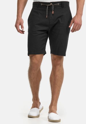 Homme portant un short noir à cordon de serrage et des chaussures blanches à enfiler, debout devant un fond blanc.