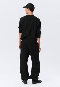 Sort langærmet skjorte og løse sorte sweatpants med sidelomme, parret med en sort cap, vist fra ryggen mod en hvid baggrund.