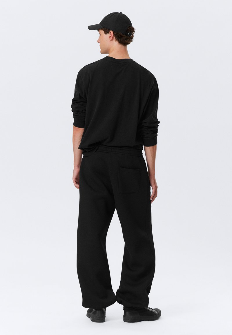 Chemise noire à manches longues et pantalon de survêtement noir ample avec poches latérales, associés à une casquette noire, présentés de dos sur fond blanc.