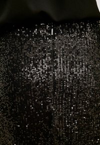 Gros plan sur un tissu noir densément recouvert de sequins noirs brillants avec une petite tirette de fermeture éclair visible en haut au centre.