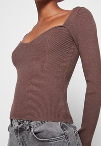 Brun ribbad långärmad topp med fyrkantig halsringning och figurnära design, matchad med grå denimjeans. Har texturerad tyg och subtila sömmar.