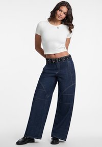 Wijde denim jeans in een donkere kleur met contrasterende stiksels, gecombineerd met een cropped wit t-shirt. Bevat een zwarte riem met metalen ringen.