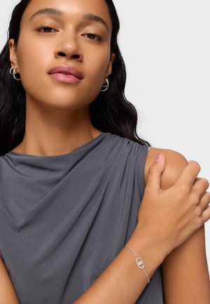 Kobieta o długich ciemnych włosach, nosząca srebrne kolczyki koła i delikatną srebrną bransoletkę, ubrana w bezrękawnik w kolorze szarym, trzymająca się za ramię.