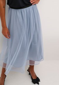 Világoskék, félig áttetsző tüll szoknya, rugalmas fekete derékkal. Az anyag lágyan omlik és finoman áramlik a mozgás során.