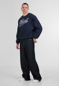 Sudadera navy con logo blanco y rojo, combinada con jeans de corte holgado en denim oscuro y zapatillas blancas, de pie sobre un fondo liso.