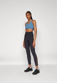 Blue sports bra with a front button design, paired with high-waisted black leggings featuring sheer side panels. Black athletic shoes.