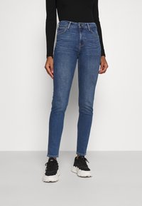 Jeans ajustados de color azul con una cintura de altura media, que presentan un diseño de cinco bolsillos, costuras sutiles y una textura de denim suave. Combinados con zapatillas negras.