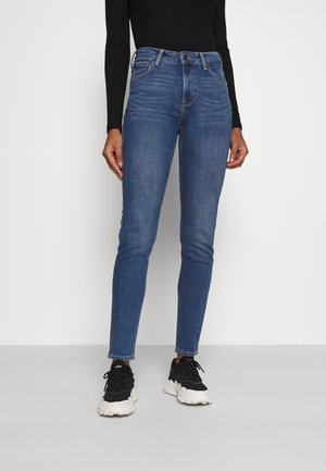 Jeans ajustados de color azul con una cintura de altura media, que presentan un diseño de cinco bolsillos, costuras sutiles y una textura de denim suave. Combinados con zapatillas negras.