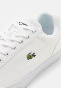 Hvite kanvas sneakers med lavt design, med en grønn krokodillelogo på siden og hvite snørebånd. Strukturerte overflater, gummisåle.
