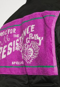 Black jacket featuring a purple terrycloth patch with white text and green accents, displaying a graphic and decorative details.