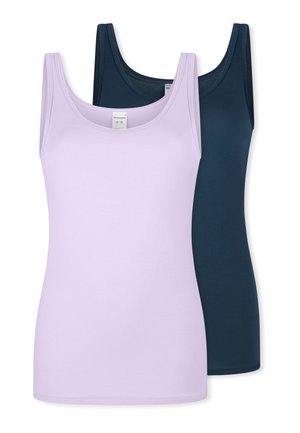 Frau trägt ein eng anliegendes hellviolettes Tanktop und passende Unterwäsche, steht vor einem einfarbigen hellen Hintergrund.