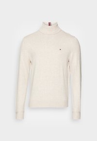 Pull à col roulé crème avec des manches longues, fabriqué en tissu doux ; présente un petit logo avec des accents rouge, blanc et bleu sur le col.