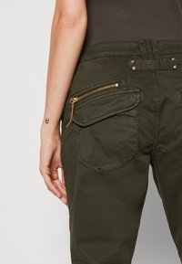 Pantalon vert foncé avec plusieurs poches arrière, dont une fermée par une fermeture éclair dorée, porté par une personne avec un petit tatouage sur le poignet gauche.