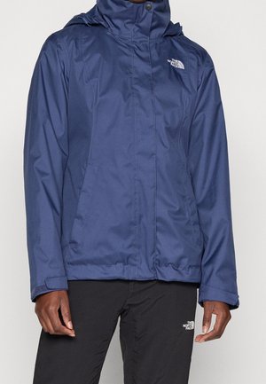 Persoon draagt een donkerblauwe capuchonjas en zwarte broek, beide met het The North Face-logo op de borst en dij.