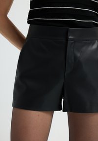 Pantaloni corti neri in ecopelle indossati con calze trasparenti nere e una maglietta a righe bianco e nera, mostrati dalla vita fino a metà coscia.