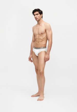 Man standing barefoot in white Emporio Armani briefs against a plain light background, facing slightly to the right.
