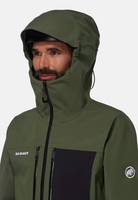 Veste verte résistante à l'eau avec capuche zippée, poche poitrine noire et logo réfléchissant. Présente un design épuré et une texture lisse.