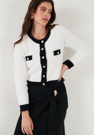 White and black cardigan with fluffy texture, gold buttons, and black trim. Features two pockets with black accents.