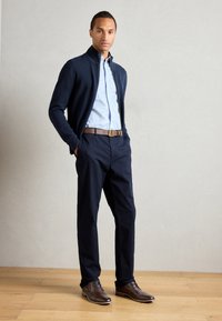 Cárdigan navy con cremallera sobre una camisa de botones azul claro, combinado con pantalones oscuros y zapatos de cuero marrón. Texturas lisas, corte clásico.