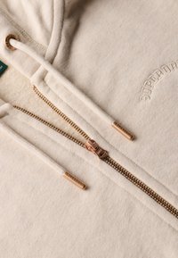 Sweatshirt à capuche couleur crème, en mélange de coton, avec une fermeture éclair à l'avant et une quincaillerie en bronze, et un logo brodé au-dessus de la fermeture.