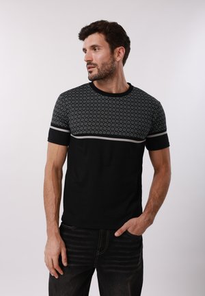 Muž v čiernych džínsoch a čiernej krátkej košeli s bielym geometrickým vzorom cez hrudník, pozerajúci sa doľava, na jednoduchom pozadí.