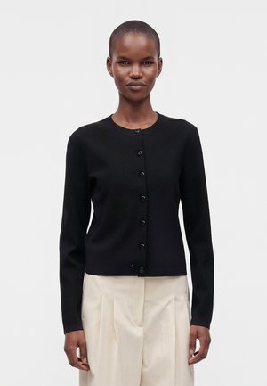 Kvinde iført en sort knap-cardigan og lyse beige plisserede bukser står mod en ensfarvet baggrund.